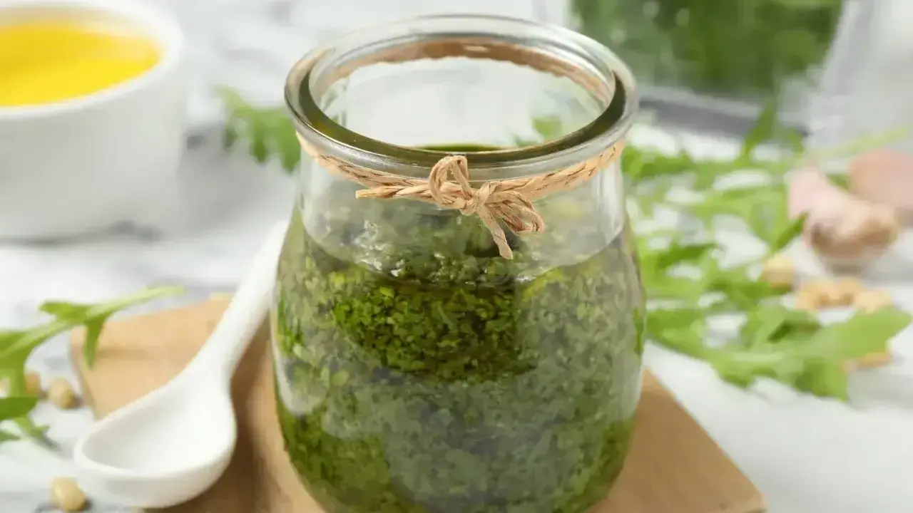 Słoik domowego pesto z rukoli, obok biała łyżeczka, piniowe orzeszki i czosnek. Idealne do makaronu!