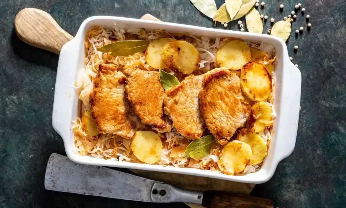 Pieczony schab w kapuście kiszonej z ziemniakami, podany w białym naczyniu, ozdobiony liśćmi laurowymi.