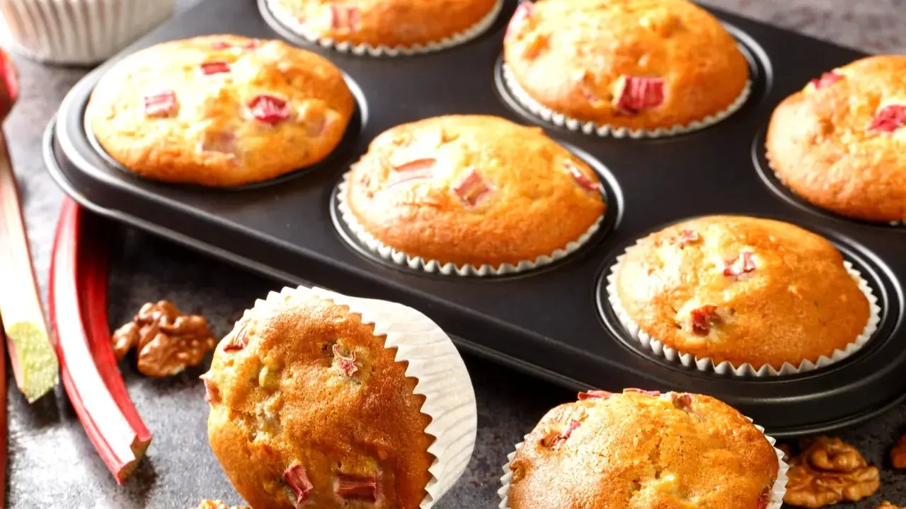Pyszne muffinki z rabarbarem, świeże i pachnące, gotowe do podania. Obok leżą łodygi rabarbaru i orzechy włoskie.