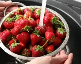 Ręce trzymają durszlak pełen soczystych truskawek, przez które przelewa się strumień wody. Tak właśnie myjemy truskawki przed jedzeniem.