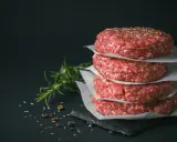 Stos świeżego mięsa na burgery, przyprawionego ziołami, obok gałązki rozmarynu i pieprzu.