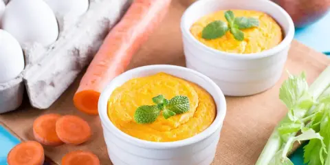 Dwa białe naczynia z pomarańczowym puree z marchewki, udekorowane miętą. Obok marchewka, jajka i seler.