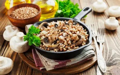 Pyszne kaszotto z pieczarkami w żeliwnej patelni, udekorowane natką pietruszki. Obok świeże pieczarki i miseczka kaszy gryczanej.