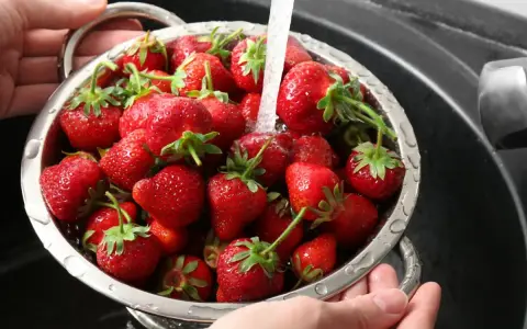 Ręce trzymają durszlak pełen soczystych truskawek, przez które przelewa się strumień wody. Tak właśnie myjemy truskawki przed jedzeniem.