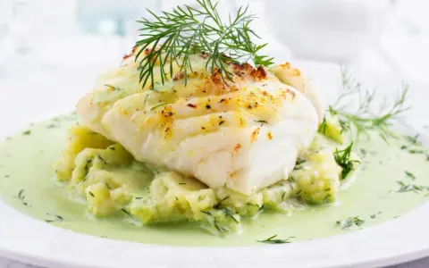 Pyszna ryba w sosie koperkowym, podana na purée ziemniaczanym z dodatkiem świeżego koperku. Danie wygląda apetycznie i aromatycznie.