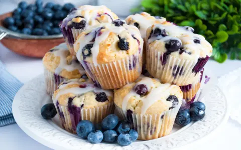 Stosik pysznych muffinek z borówkami, polanych lukrem. Obok świeże borówki i zielone listki.