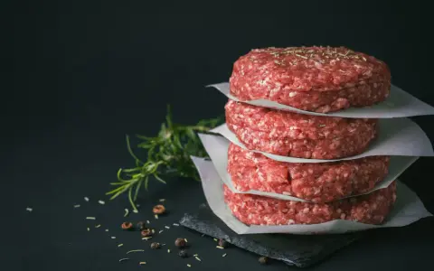Stos świeżego mięsa na burgery, przyprawionego ziołami, obok gałązki rozmarynu i pieprzu.