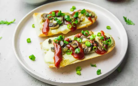 Dwie zapiekanki z pieczarkami, serem i keczupem, posypane szczypiorkiem, na talerzu.