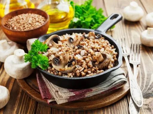 Pyszne kaszotto z pieczarkami w żeliwnej patelni, udekorowane natką pietruszki. Obok świeże pieczarki i miseczka kaszy gryczanej.