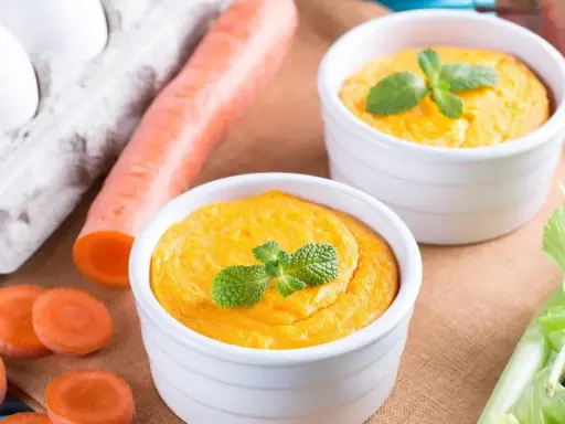 Dwa białe naczynia z pomarańczowym puree z marchewki, udekorowane miętą. Obok marchewka, jajka i seler.