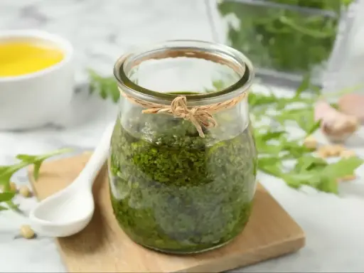 Słoik domowego pesto z rukoli, obok biała łyżeczka, piniowe orzeszki i czosnek. Idealne do makaronu!