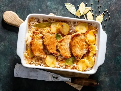 Pieczony schab w kapuście kiszonej z ziemniakami, podany w białym naczyniu, ozdobiony liśćmi laurowymi.