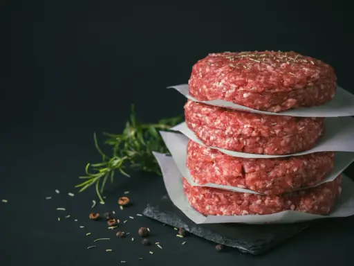 Stos świeżego mięsa na burgery, przyprawionego ziołami, obok gałązki rozmarynu i pieprzu.