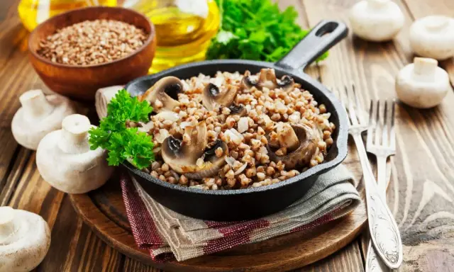 Pyszne kaszotto z pieczarkami w żeliwnej patelni, udekorowane natką pietruszki. Obok świeże pieczarki i miseczka kaszy gryczanej.