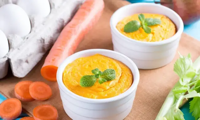 Dwa białe naczynia z pomarańczowym puree z marchewki, udekorowane miętą. Obok marchewka, jajka i seler.
