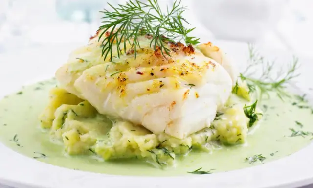 Pyszna ryba w sosie koperkowym, podana na purée ziemniaczanym z dodatkiem świeżego koperku. Danie wygląda apetycznie i aromatycznie.