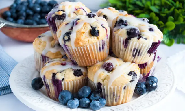Stosik pysznych muffinek z borówkami, polanych lukrem. Obok świeże borówki i zielone listki.