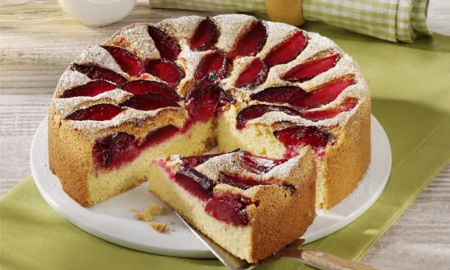 Pyszne ciasto ze śliwkami, posypane cukrem pudrem, z kawałkiem na łopatce. Obok miseczka bitej śmietany.