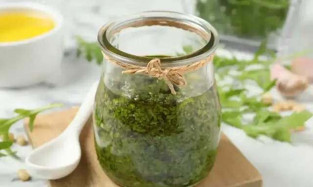 Słoik domowego pesto z rukoli, obok biała łyżeczka, piniowe orzeszki i czosnek. Idealne do makaronu!