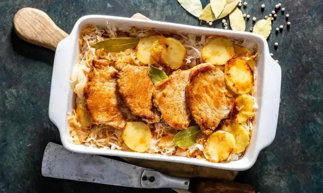 Pieczony schab w kapuście kiszonej z ziemniakami, podany w białym naczyniu, ozdobiony liśćmi laurowymi.