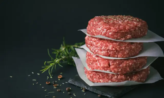 Stos świeżego mięsa na burgery, przyprawionego ziołami, obok gałązki rozmarynu i pieprzu.