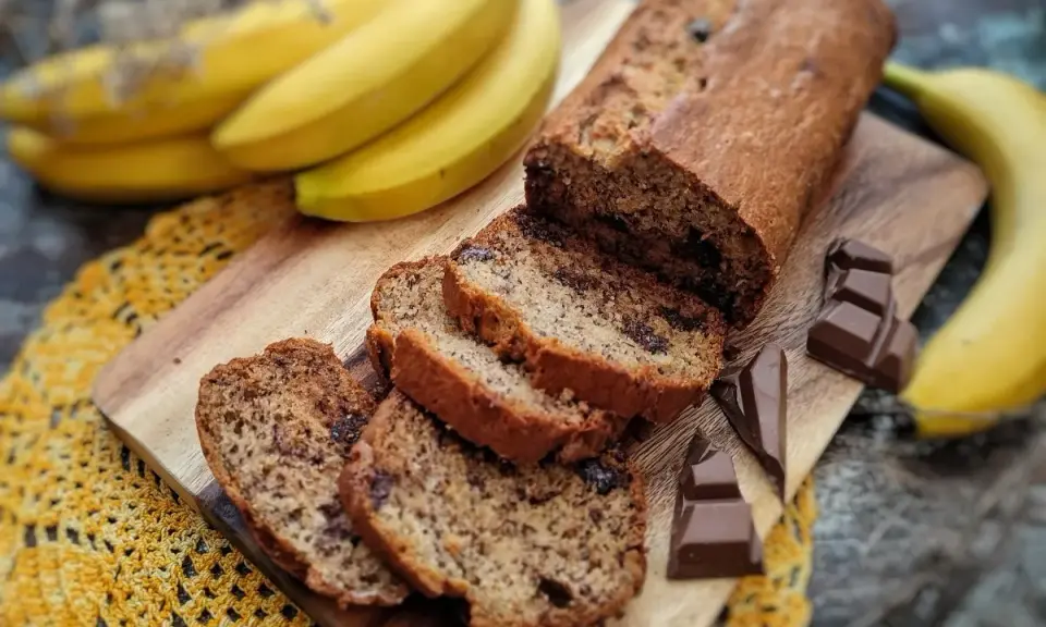 Pyszny chlebek bananowy z czekoladą, pokrojony w plastry, obok świeżych bananów i kostek czekolady.