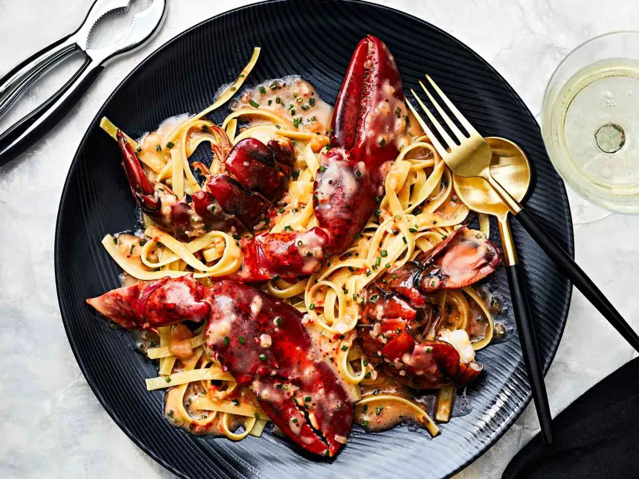 Elegancka kolacja dla dwojga: makaron linguine z homarem w kremowym sosie, podany na czarnym talerzu. Obok kieliszek białego wina.