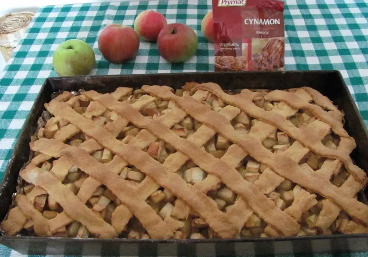 Upieczona babcina szarlotka najlepsza, pachnąca cynamonem, z jabłkami i kratką z ciasta. Obok leżą świeże jabłka.