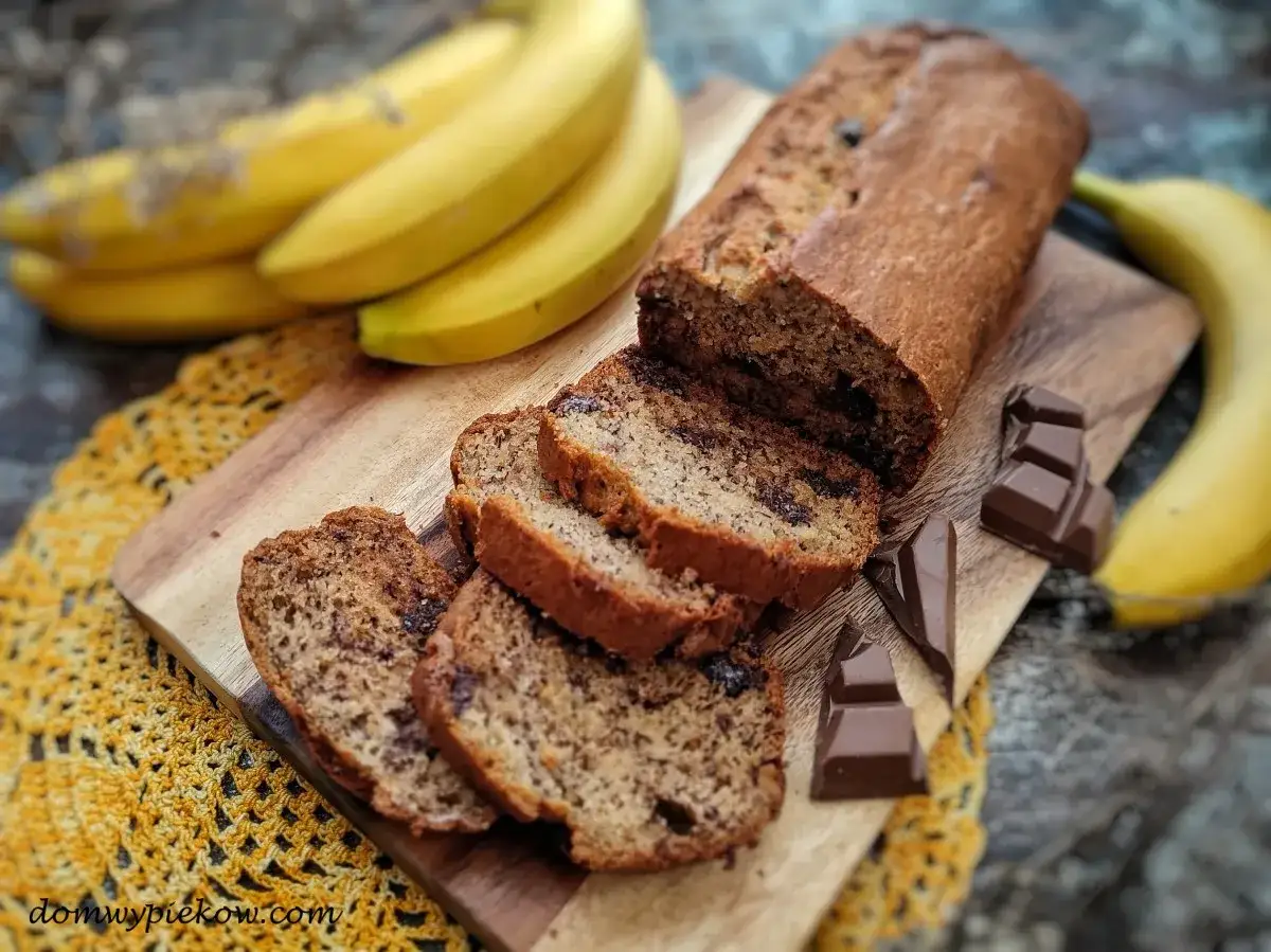 Pyszny chlebek bananowy z czekoladą, pokrojony w plastry, obok świeżych banan&oacute;w i kostek czekolady.