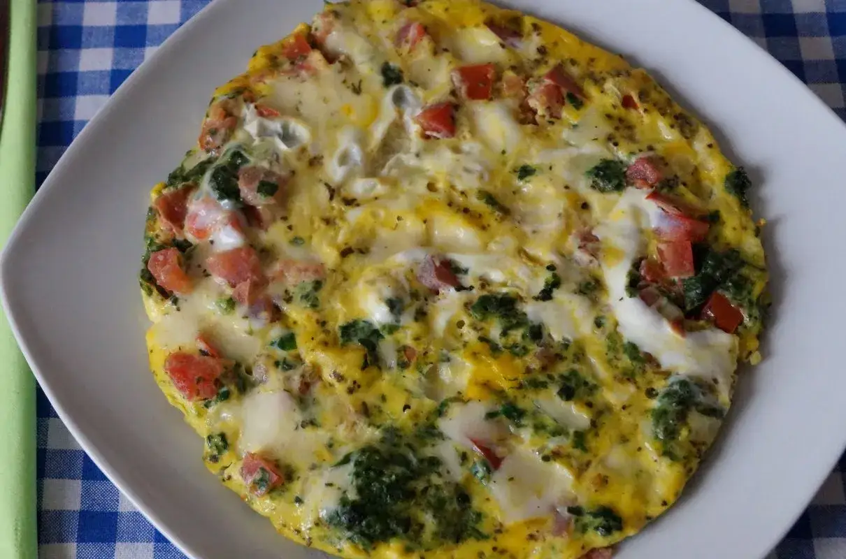 Puszysty omlet ze szpinakiem i pomidorami, podany na białym talerzu, z niebiesko-białą kratką w tle.