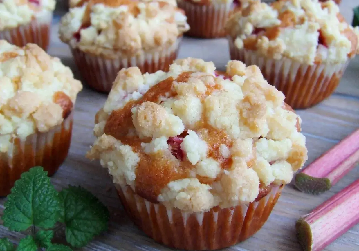 Pyszne muffinki z rabarbarem i kruszonką, pachnące latem. Obok leżą łodygi rabarbaru i listki mięty.